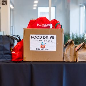 Bright orange cardboard box with "Food Drive Donates Here" sign, surrounded by reusable bags at a community food donation event.